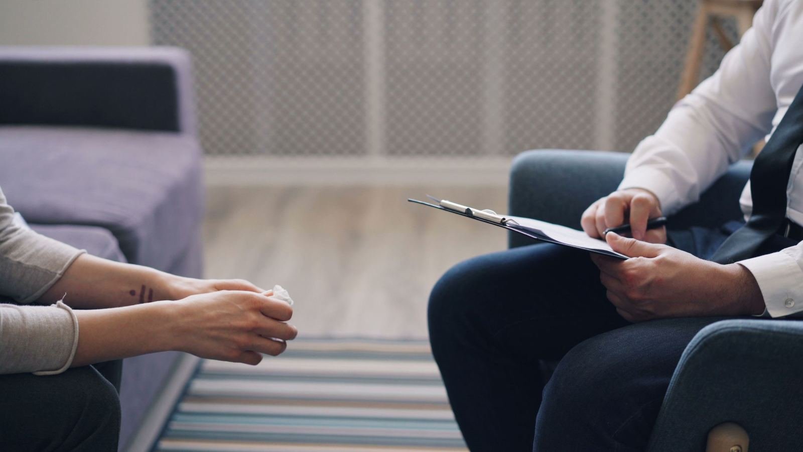 A clinician holding a clipboard sits across from someone on a couch.