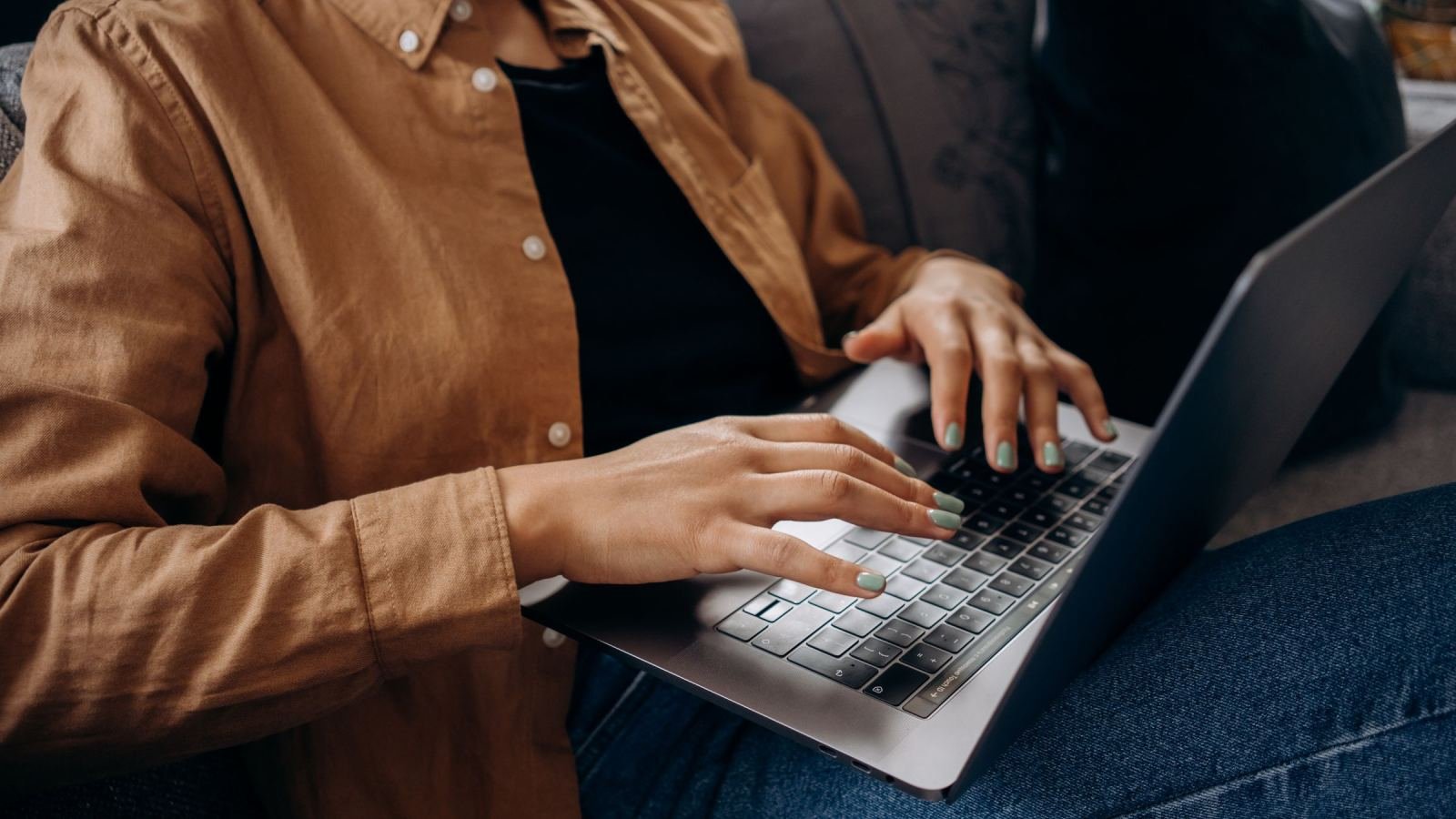 A person in a brown shirt typing on a laptop while sitting on a couch