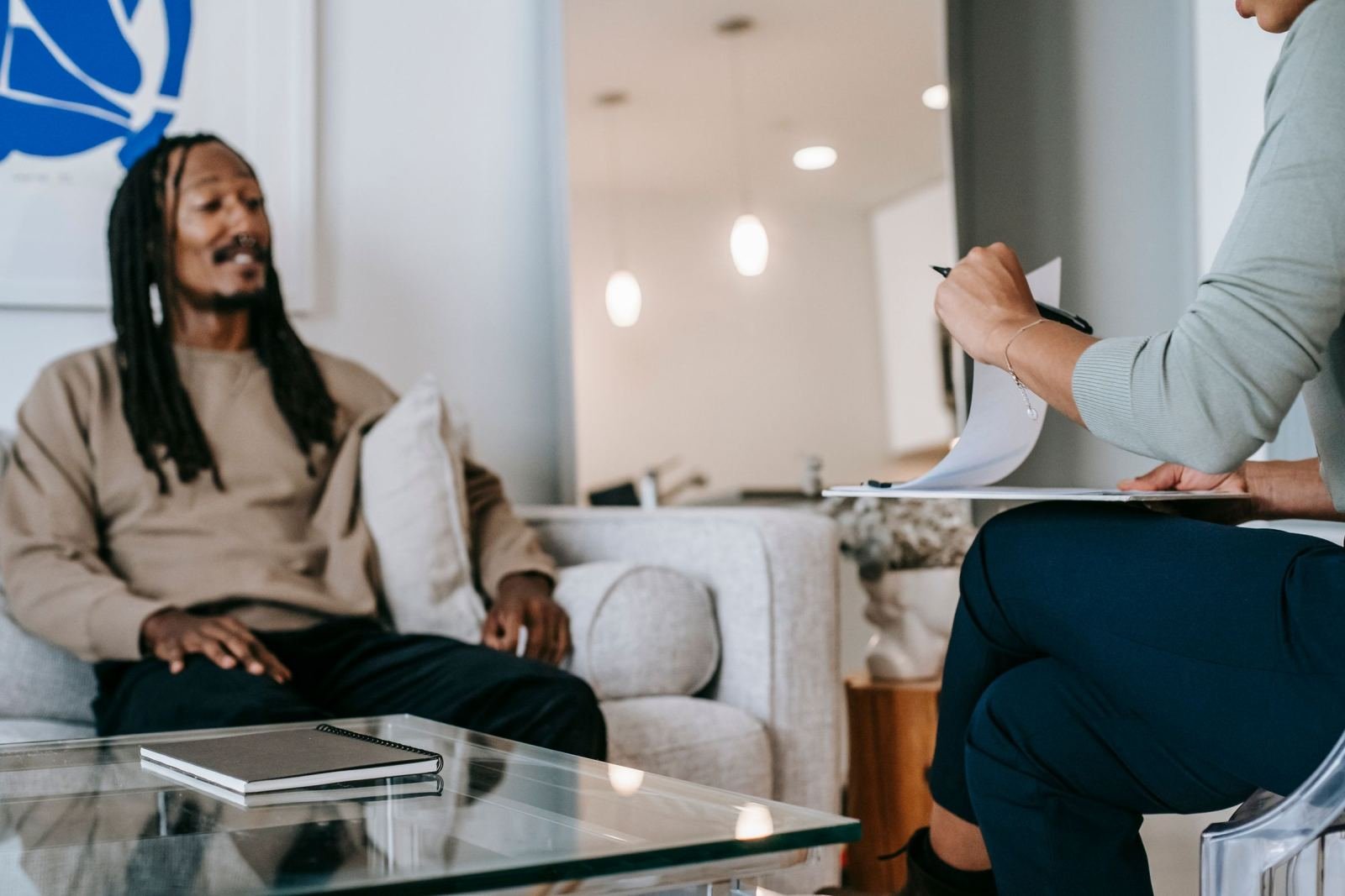 A man sits on a couch while a clinician holds a clipboard and talks with him.
