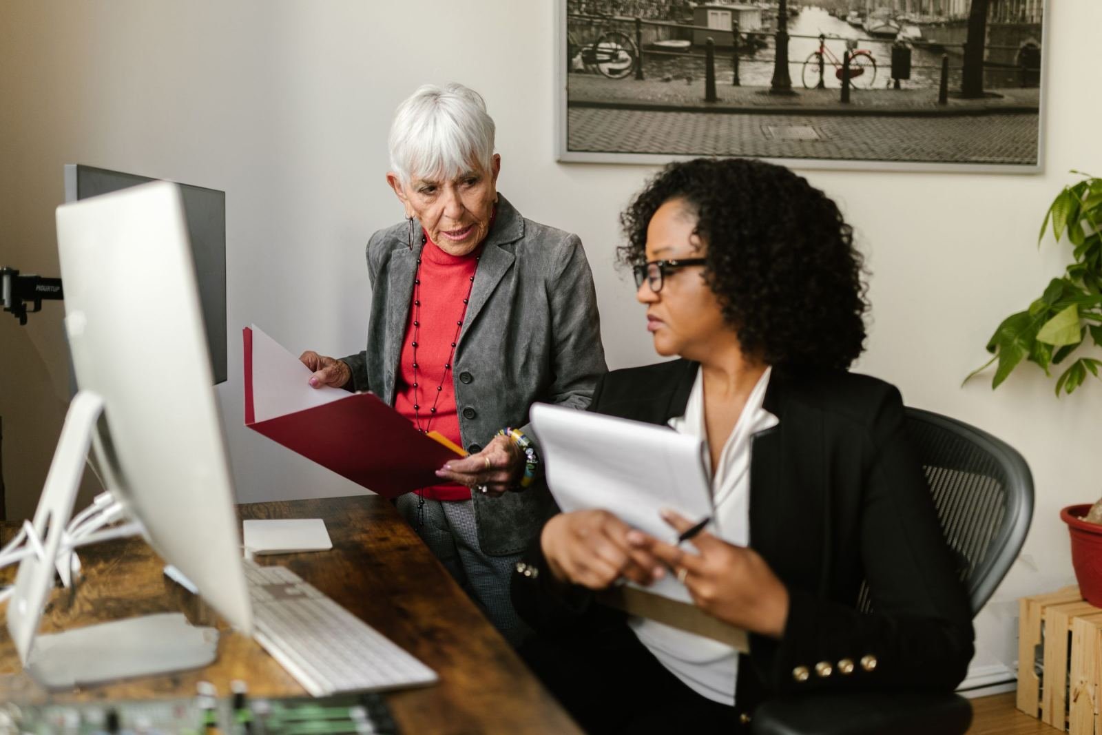 In a bright office, a seated woman listens to an older woman holding a red folder.