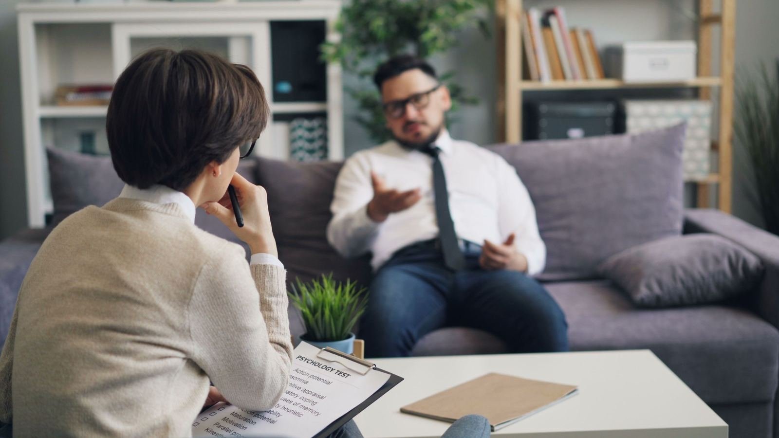 In a bright office, a clinician holds a clipboard while talking to a man in a white shirt.