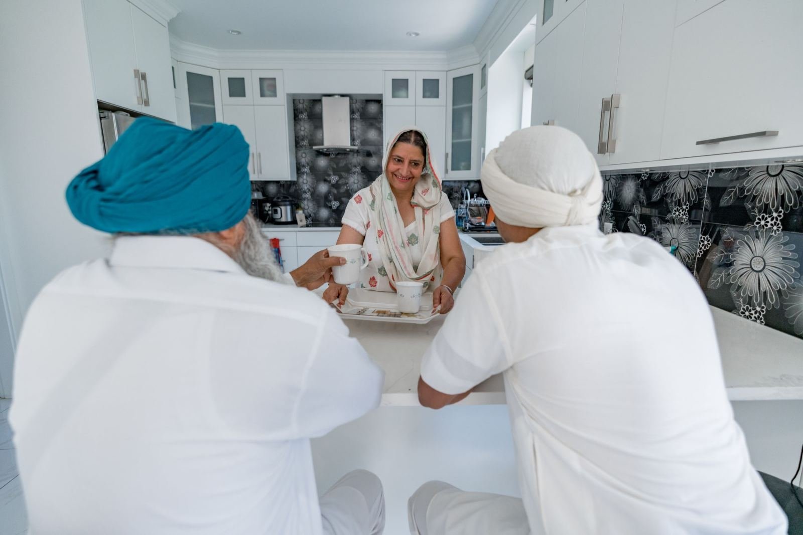 A smiling woman holds a tray of mugs. Two men in turbans sit at the kitchen counter, facing her.