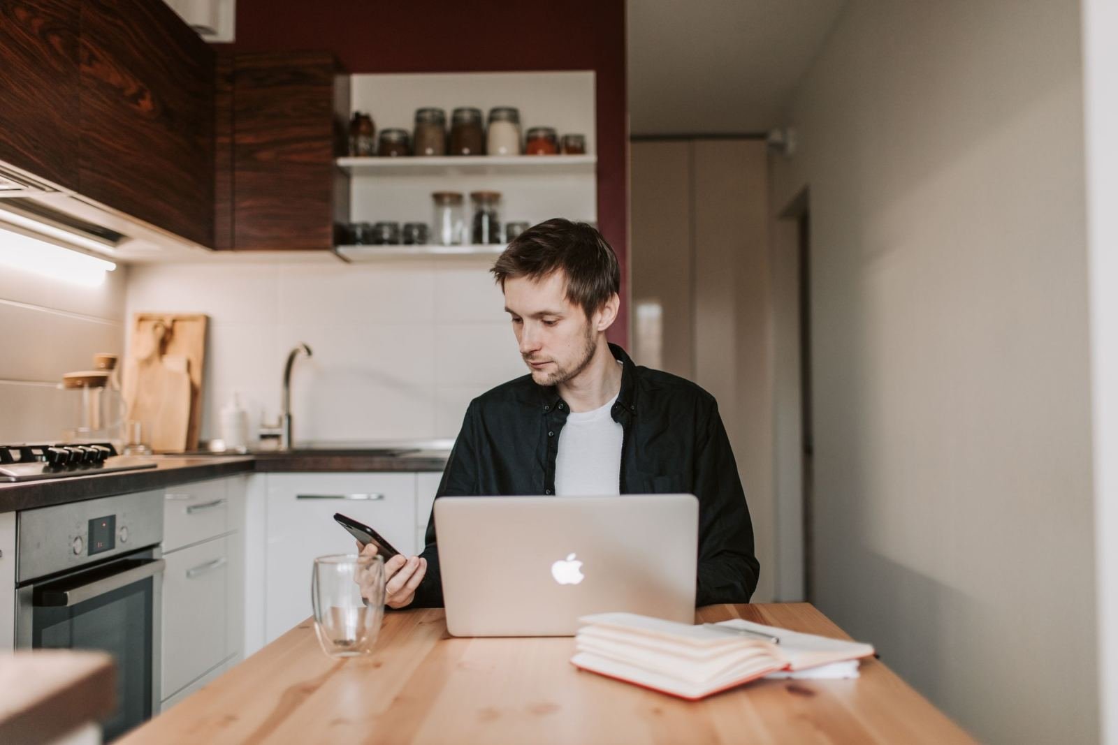A man looks at his phone while sitting at a table with a laptop and notebook