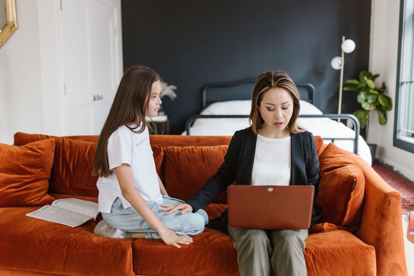 A mother and her daughter sit on an orange couch, looking at a laptop screen side by side.
