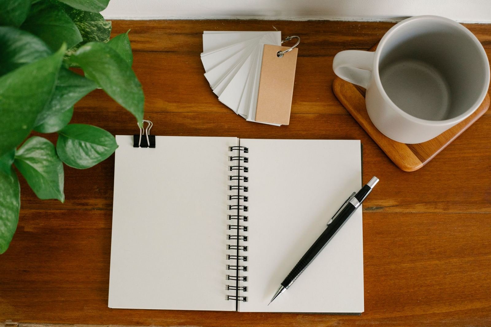 An open blank notebook with a pen, a white mug, and a stack of cards on a wooden desk