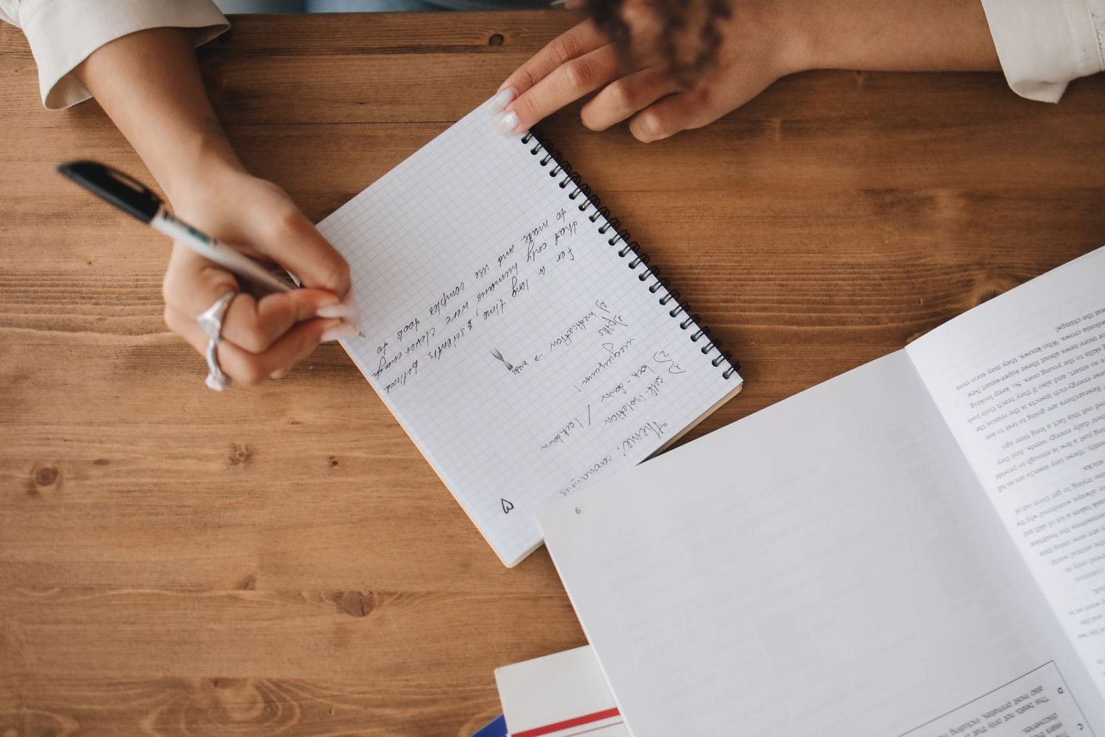 A person fills out a spiral notebook while sitting at a wooden table.