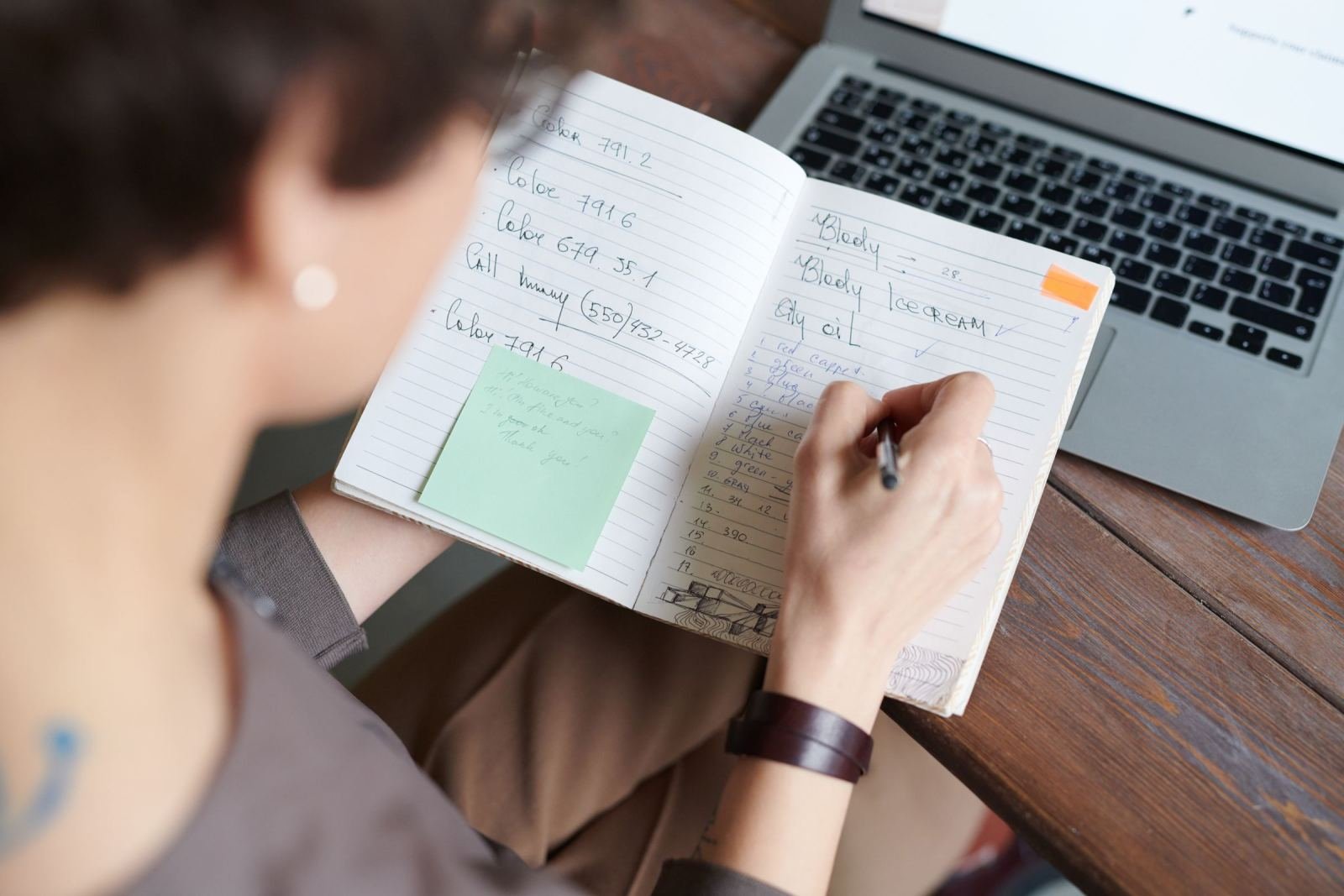 A person writing in a notebook beside an open laptop on a wooden table