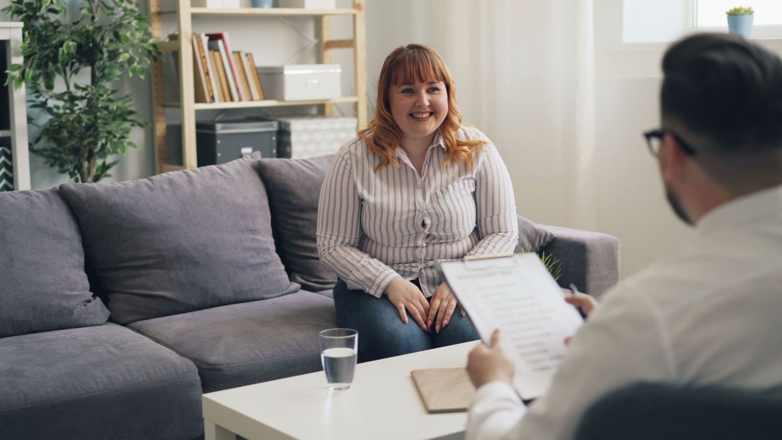A smiling woman chats with a professional who holds a clipboard in an office.