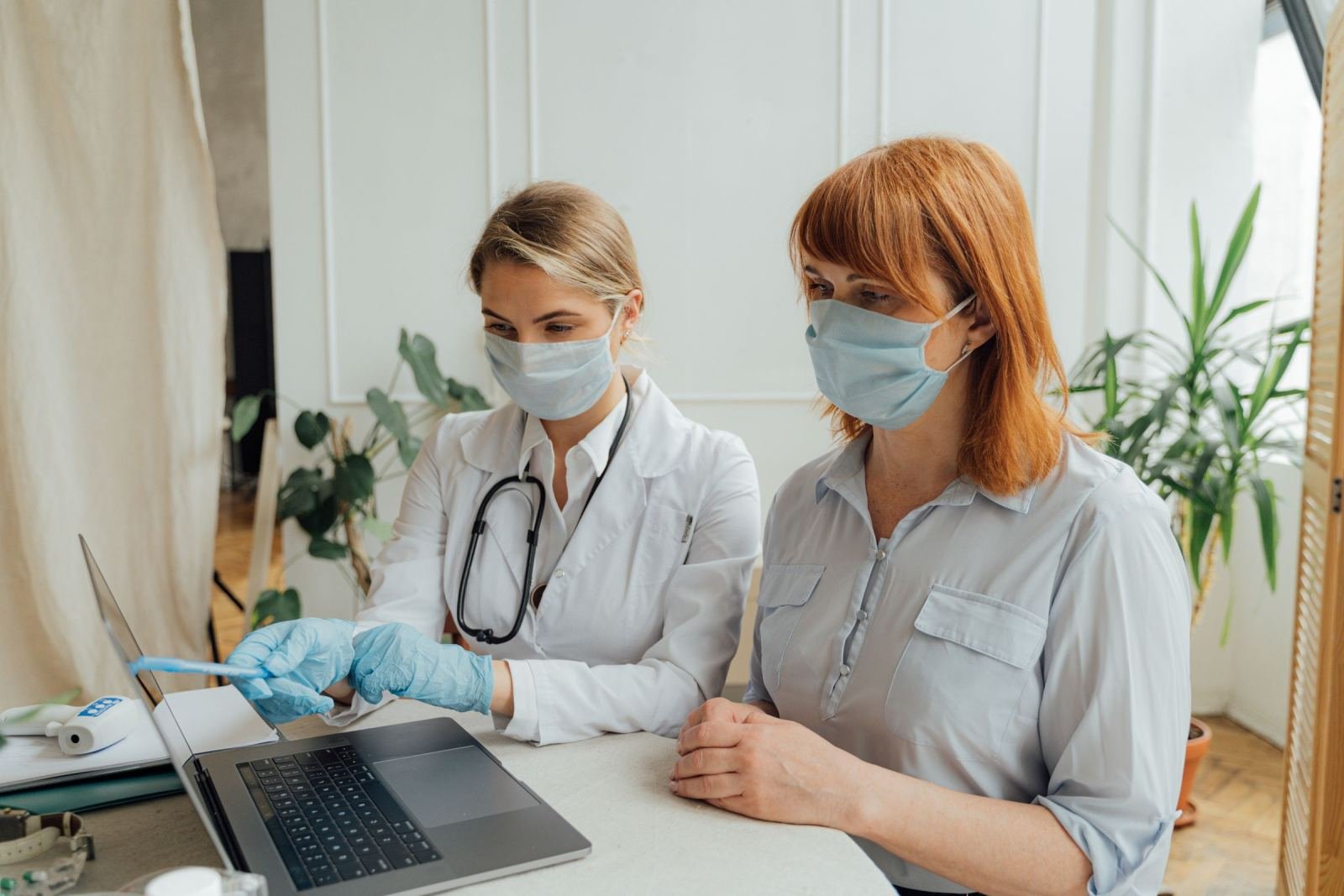 A masked clinician in a white coat shows a patient something on a laptop screen.