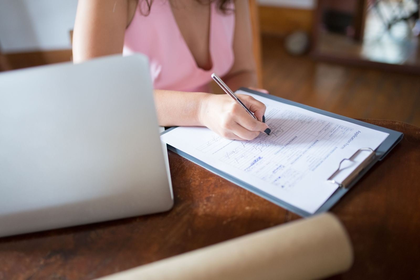 A person writing on an application form on a clipboard next to a laptop