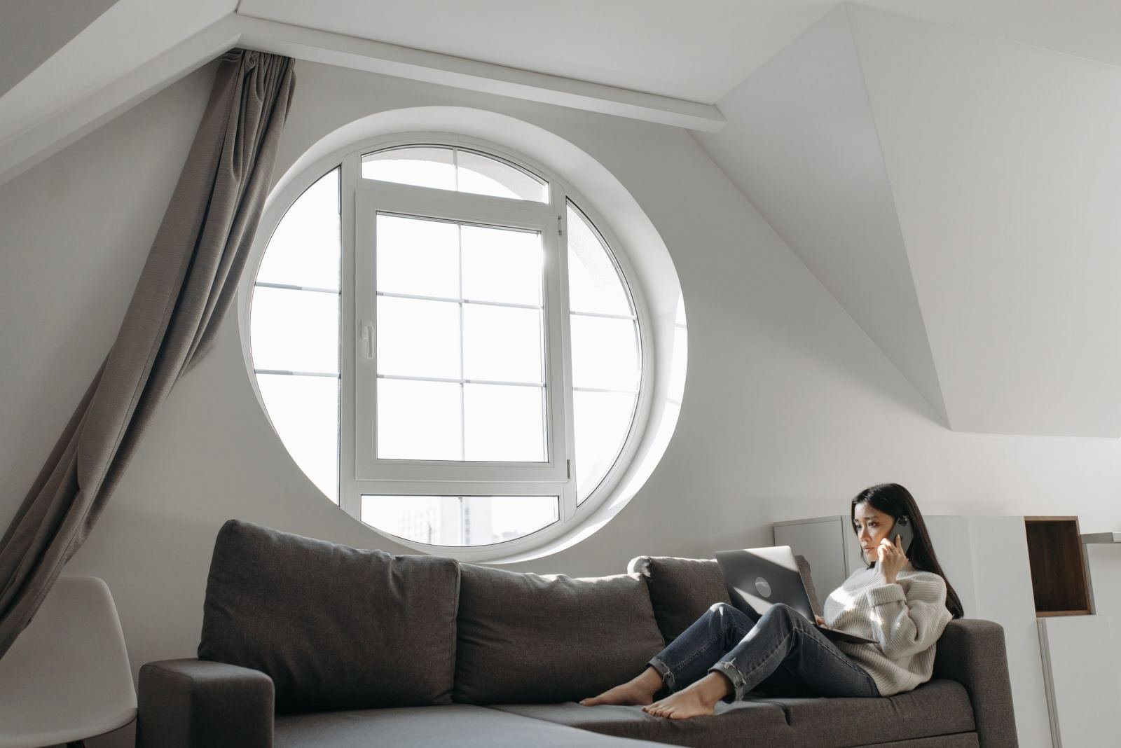 A woman sits on a grey sofa. She uses a laptop and talks on a phone.
