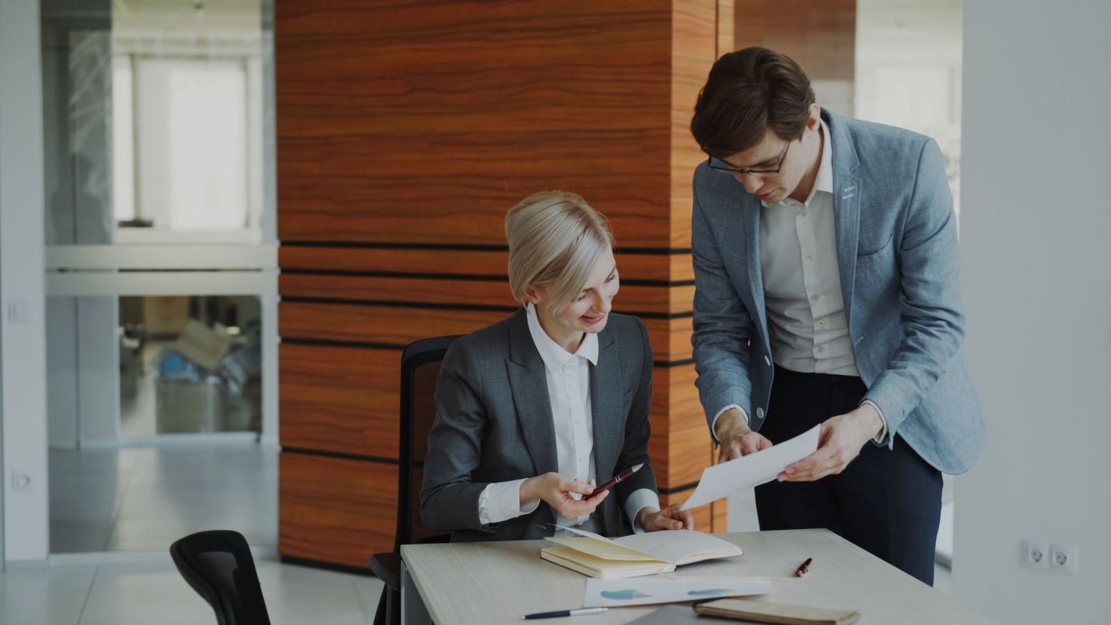 Two people in business clothes look over papers together in a well-lit office.