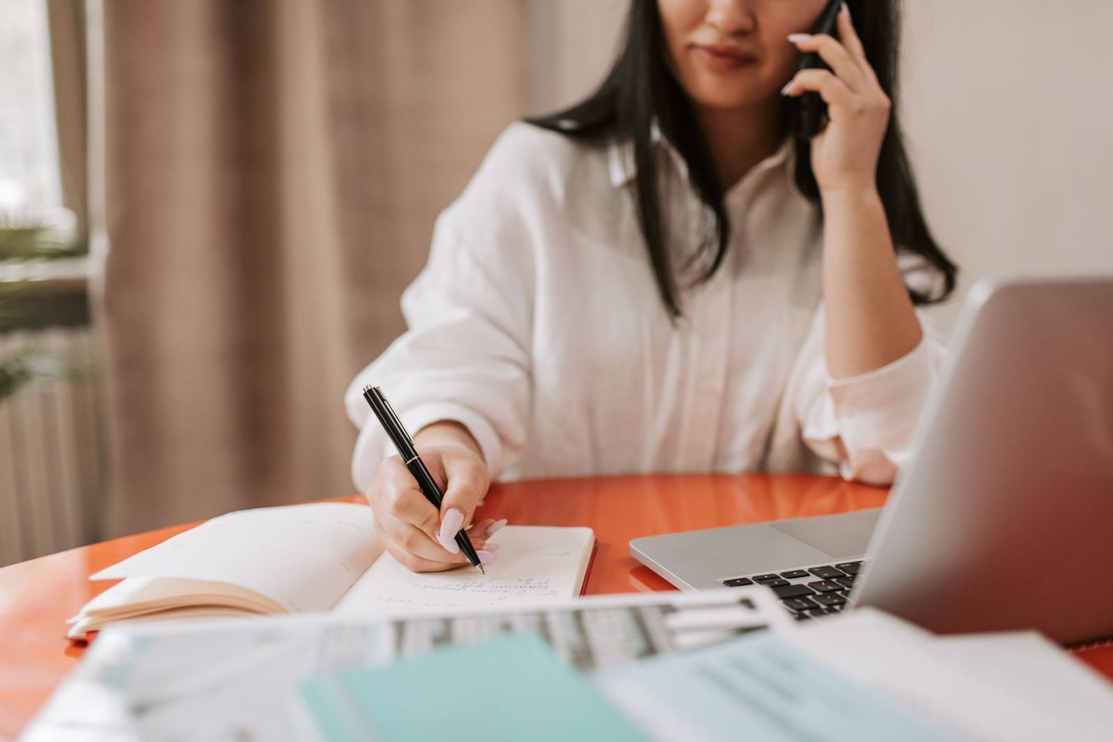 A woman uses a laptop on her desk. She holds a phone to her ear and writes in a notebook.