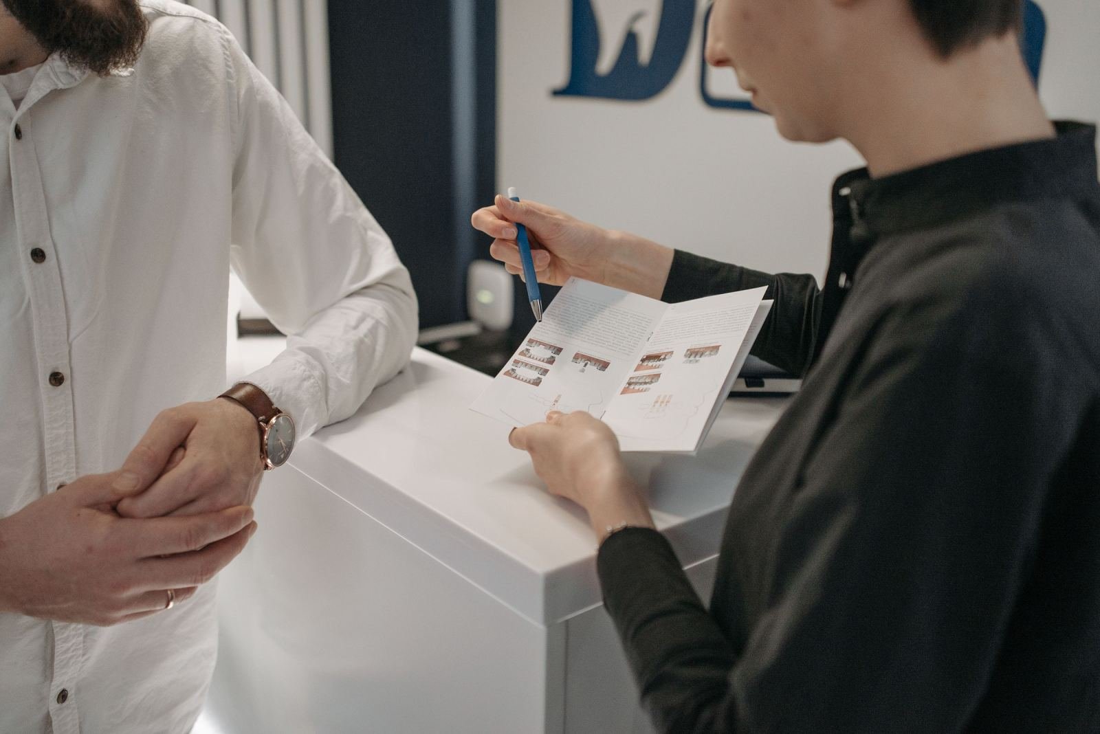 A person points to a brochure while talking to a man across a white counter