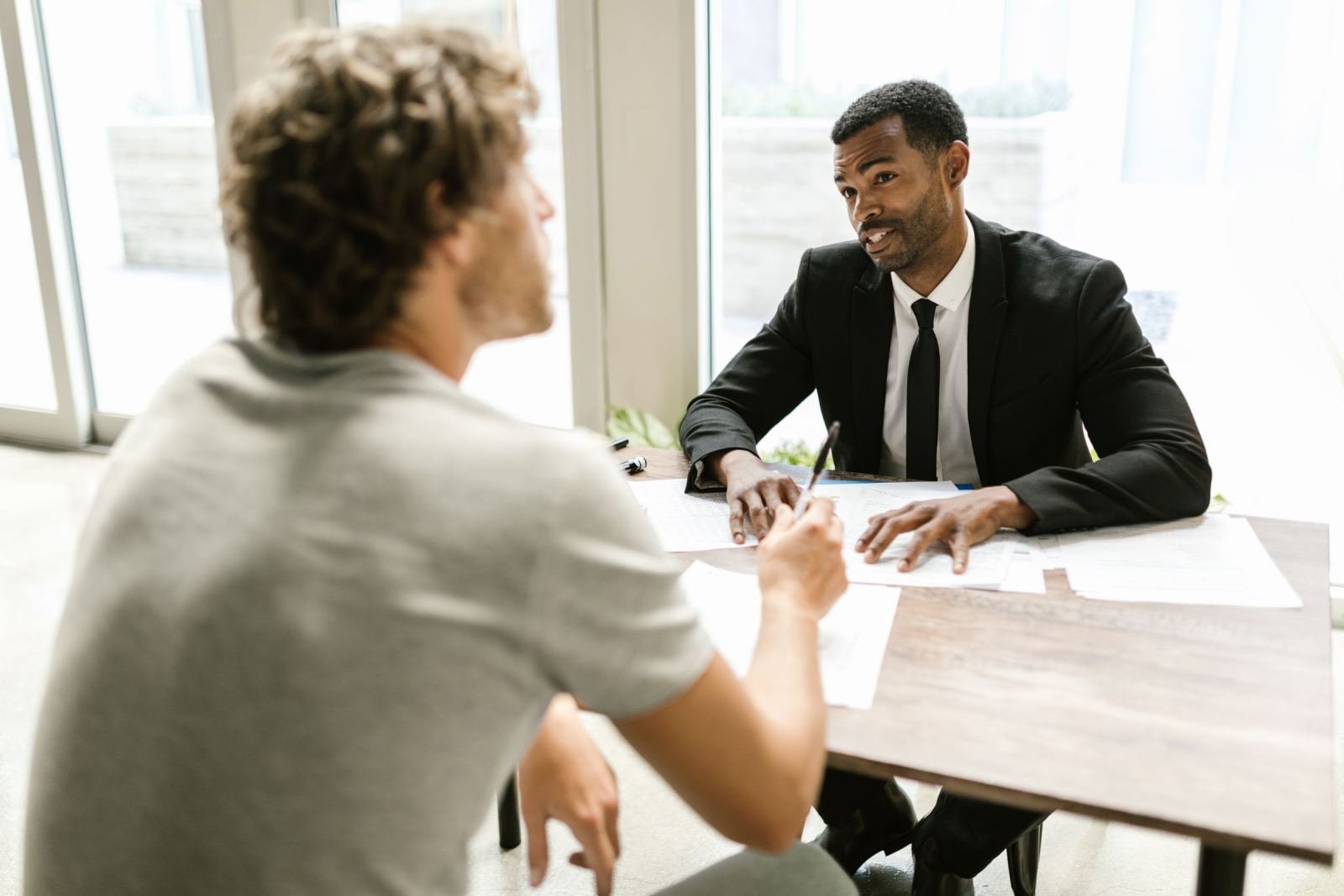 A client sits at a wooden table with a man in a suit reviewing paperwork.