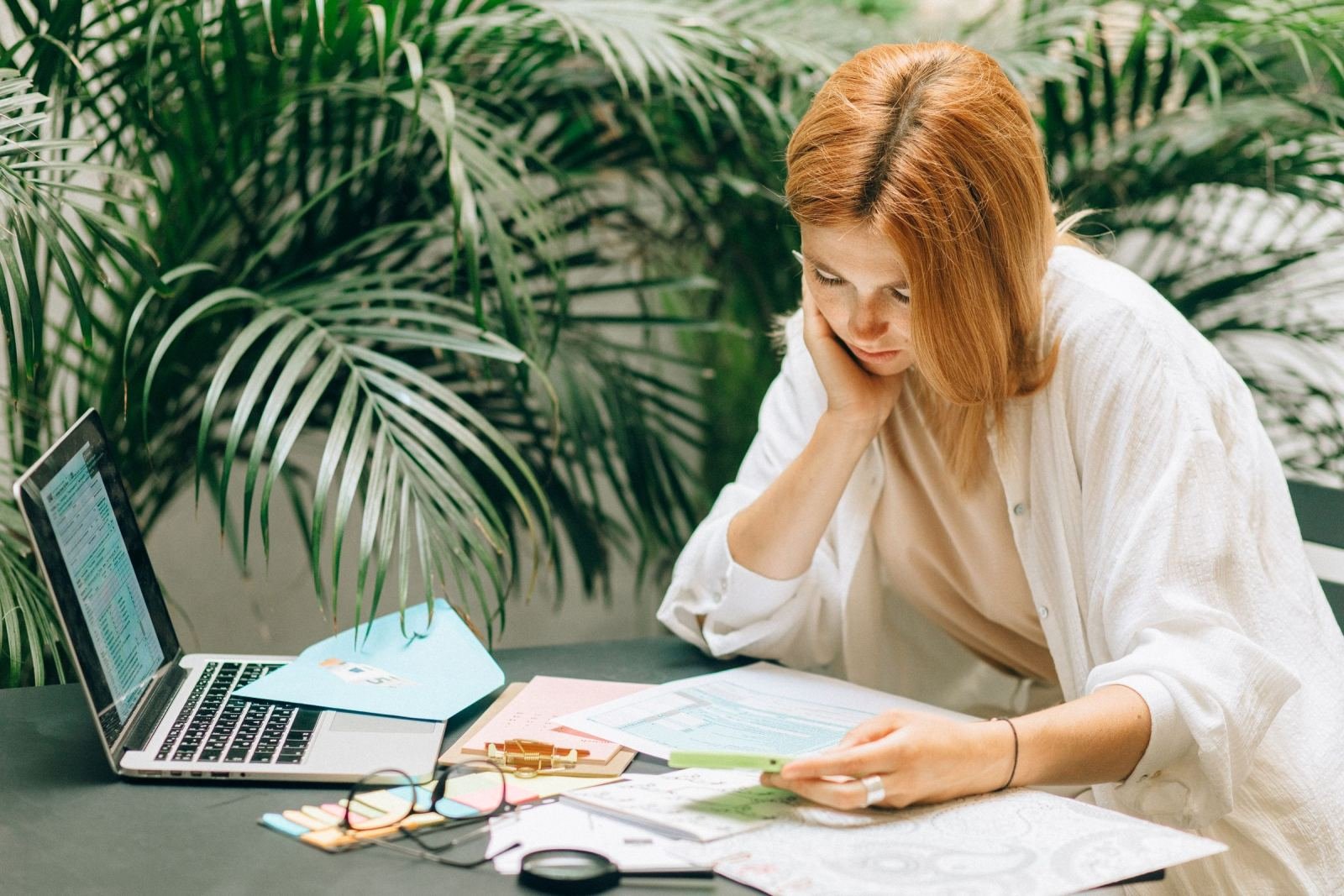 A woman looks at documents on a table next to a laptop and green plants