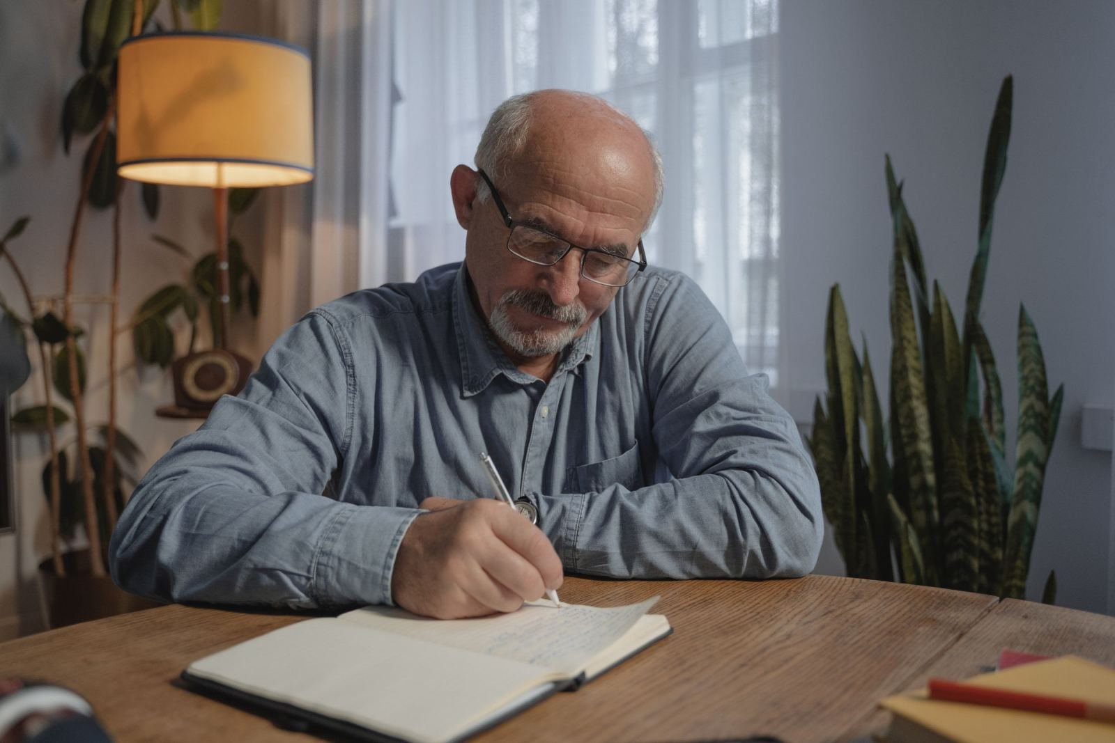 A senior man wearing glasses fills out a notebook while sitting at a wood table.