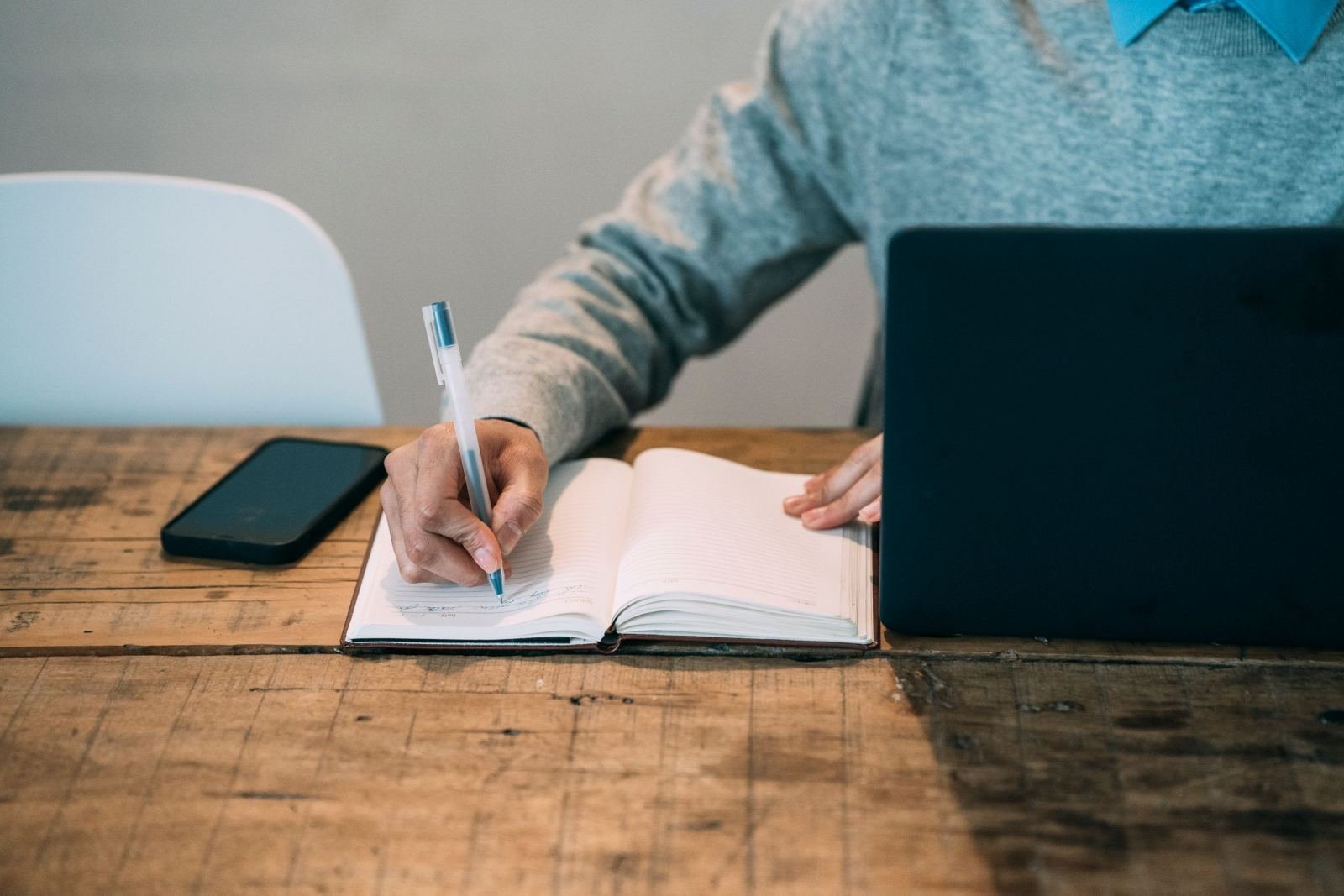 A person writes in a notebook while using a laptop and phone on a wood desk