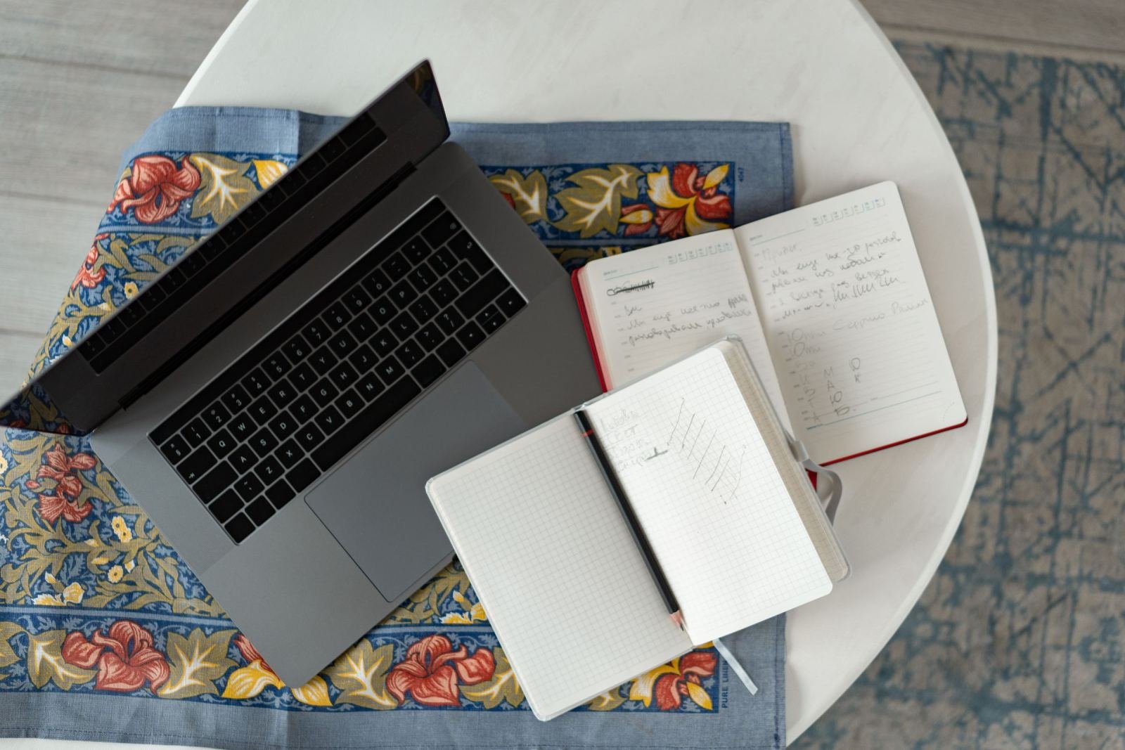 A laptop and two open notebooks with handwritten notes on a floral cloth