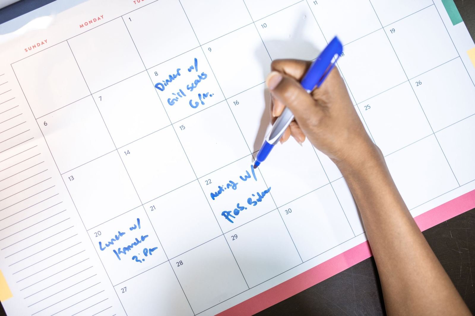 A hand using a blue pen to write notes on a paper wall calendar