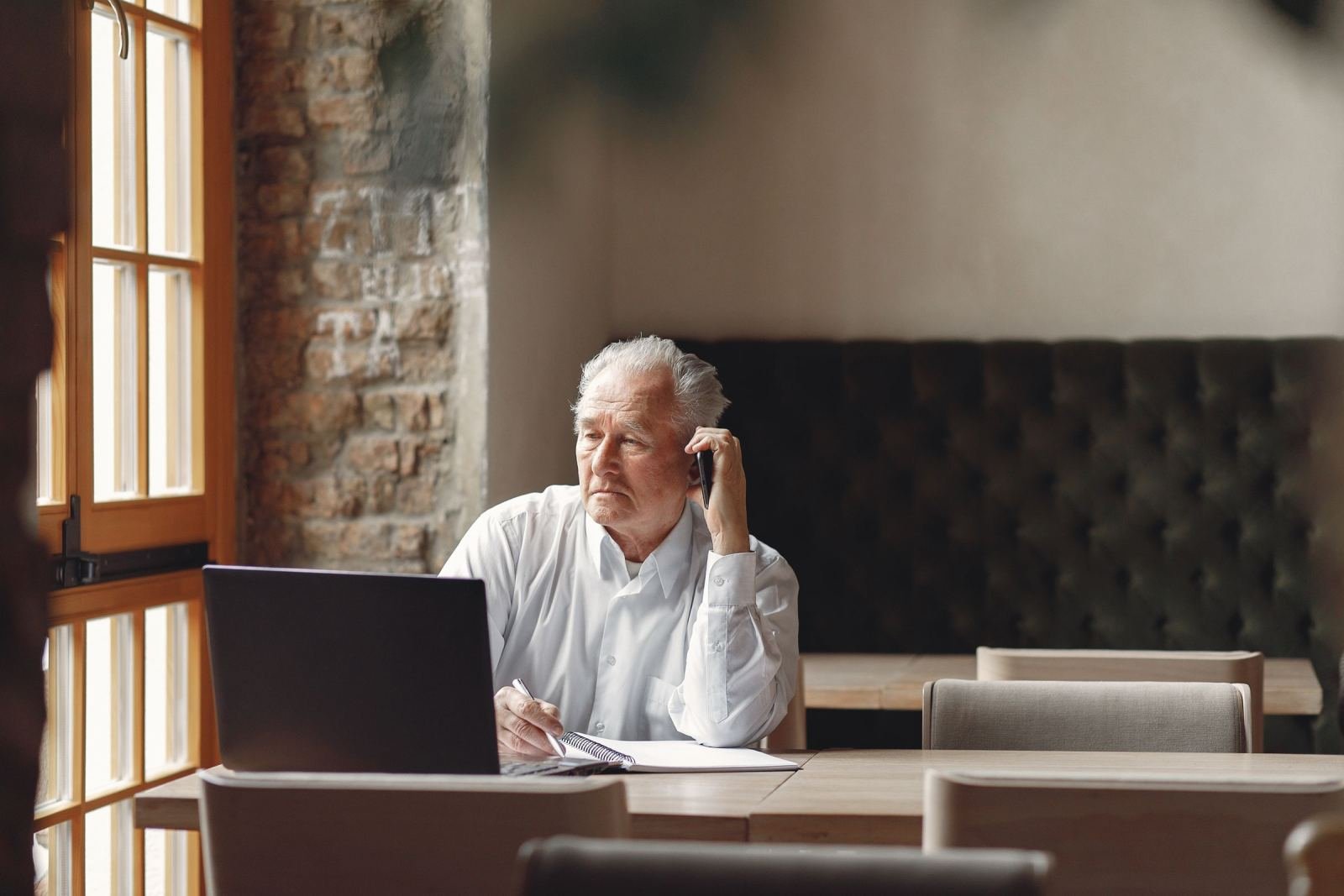 A laptop and notebook sit on a table where an older man makes a phone call