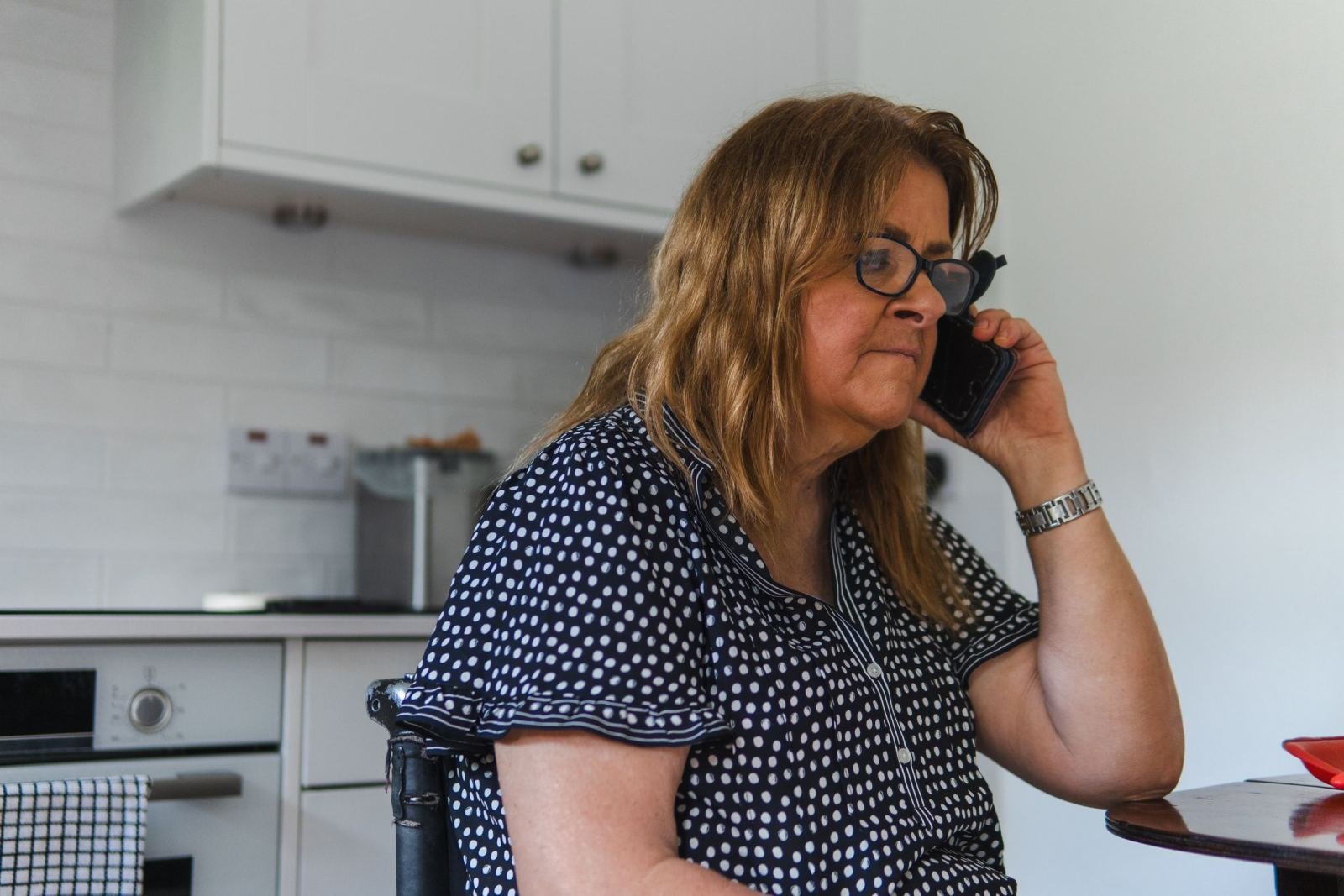 A woman wearing glasses makes a call on her phone while standing in her kitchen.