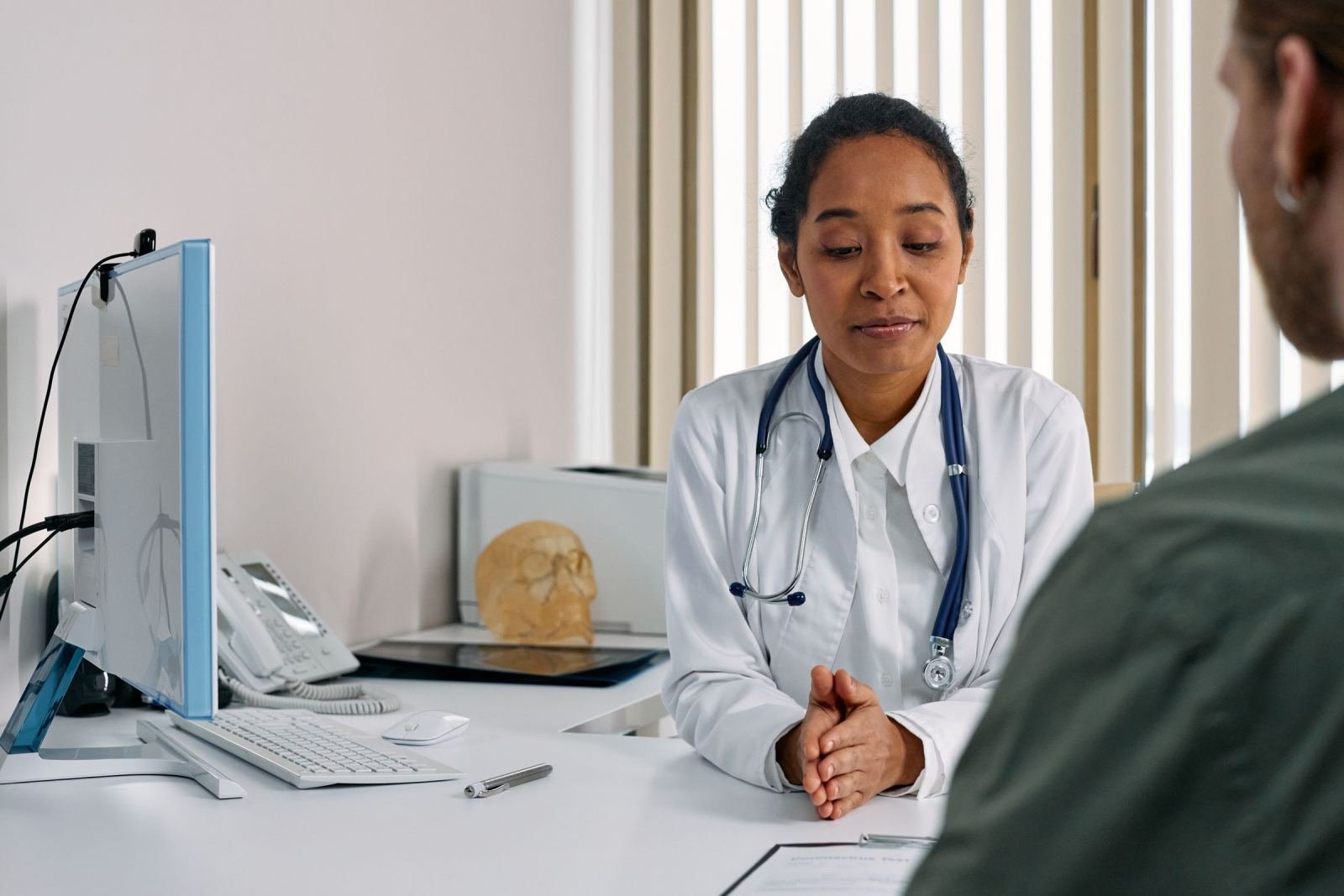 A doctor in a white coat and stethoscope speaks with a patient in a bright room