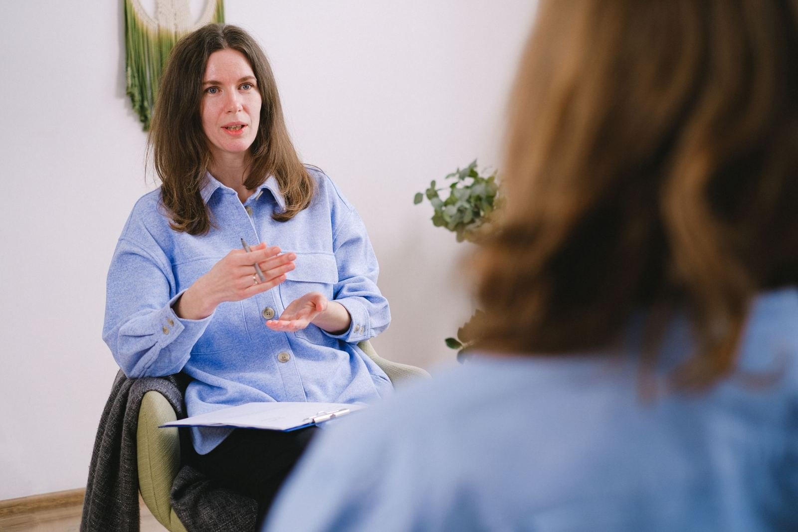 A woman in a blue shirt gestures while speaking to another person in a bright room