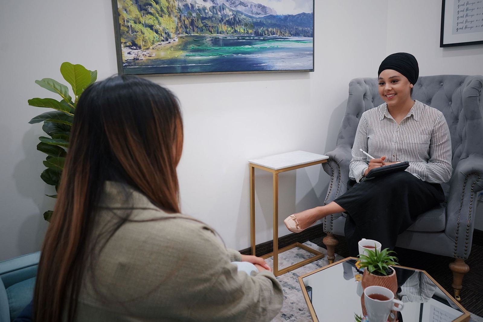 A woman in a headscarf sits in a grey armchair and smiles while talking to another person