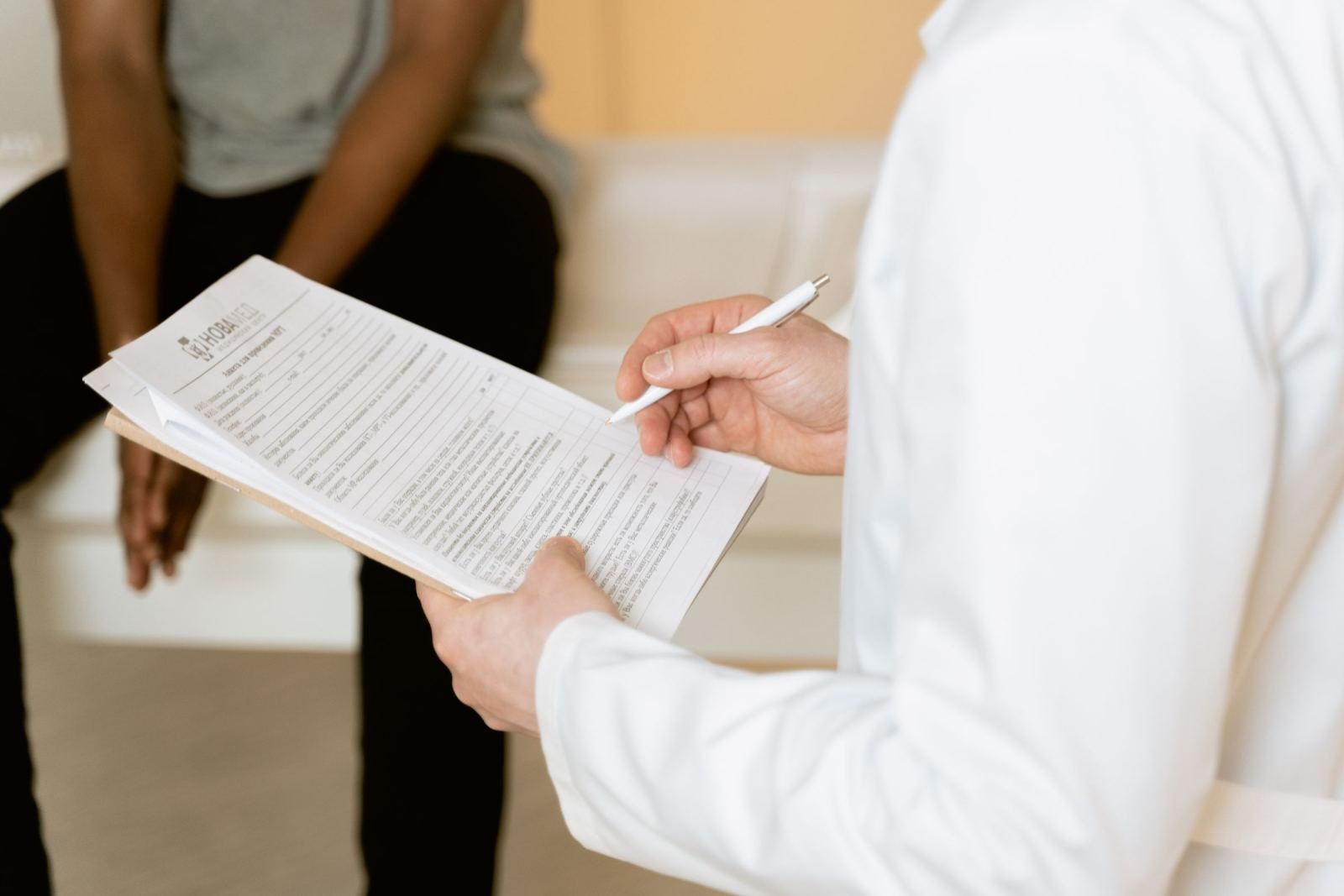 A clinician wearing a white coat marks a clipboard while a patient waits behind them