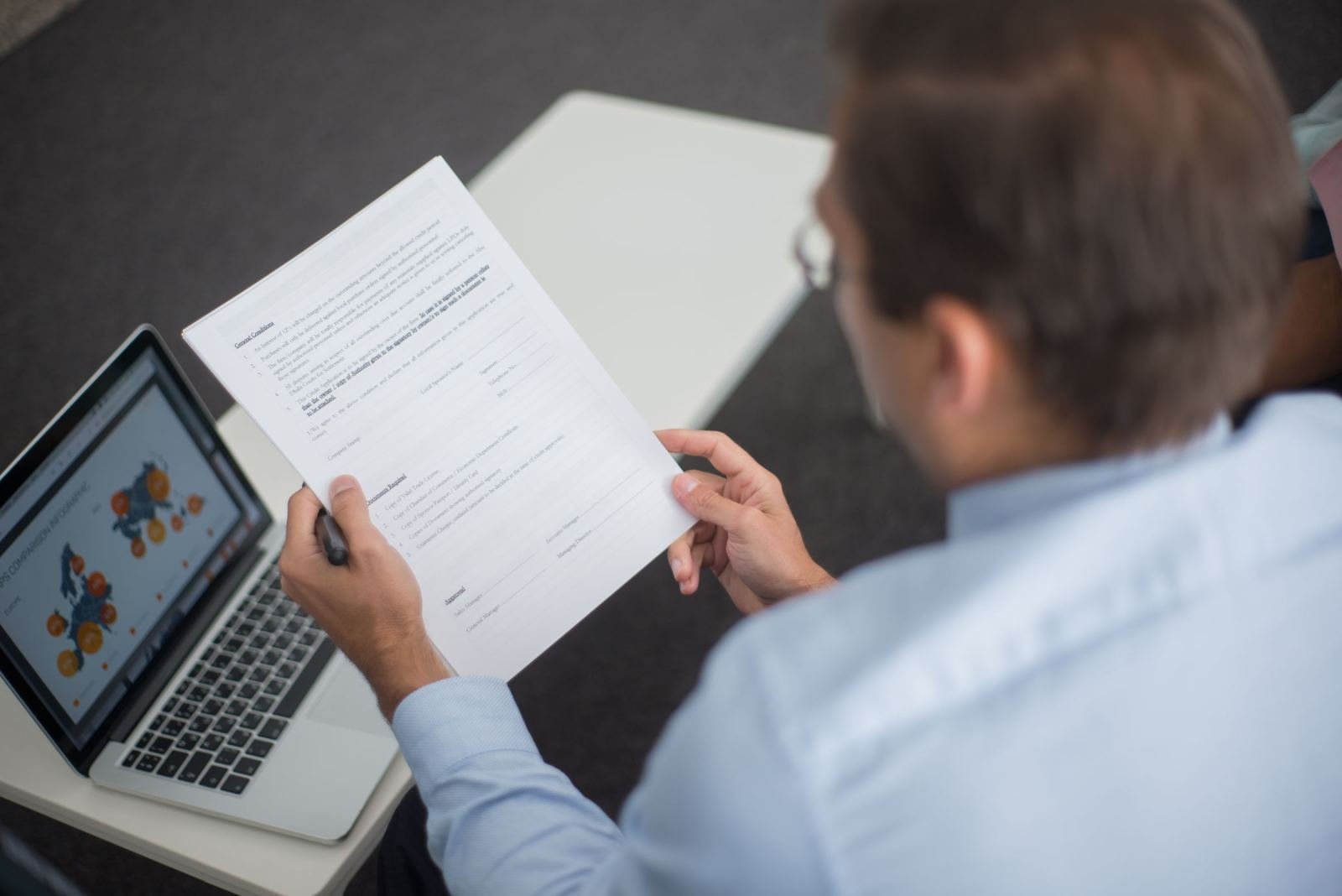 A man holds a printed document while sitting next to a laptop with a chart
