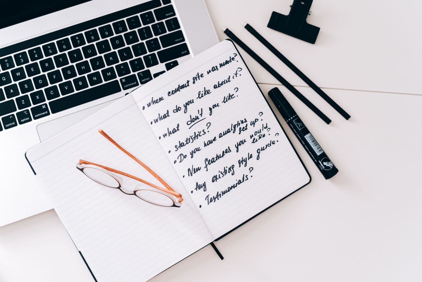 A handwritten list of questions sits in a notebook beside a laptop and glasses