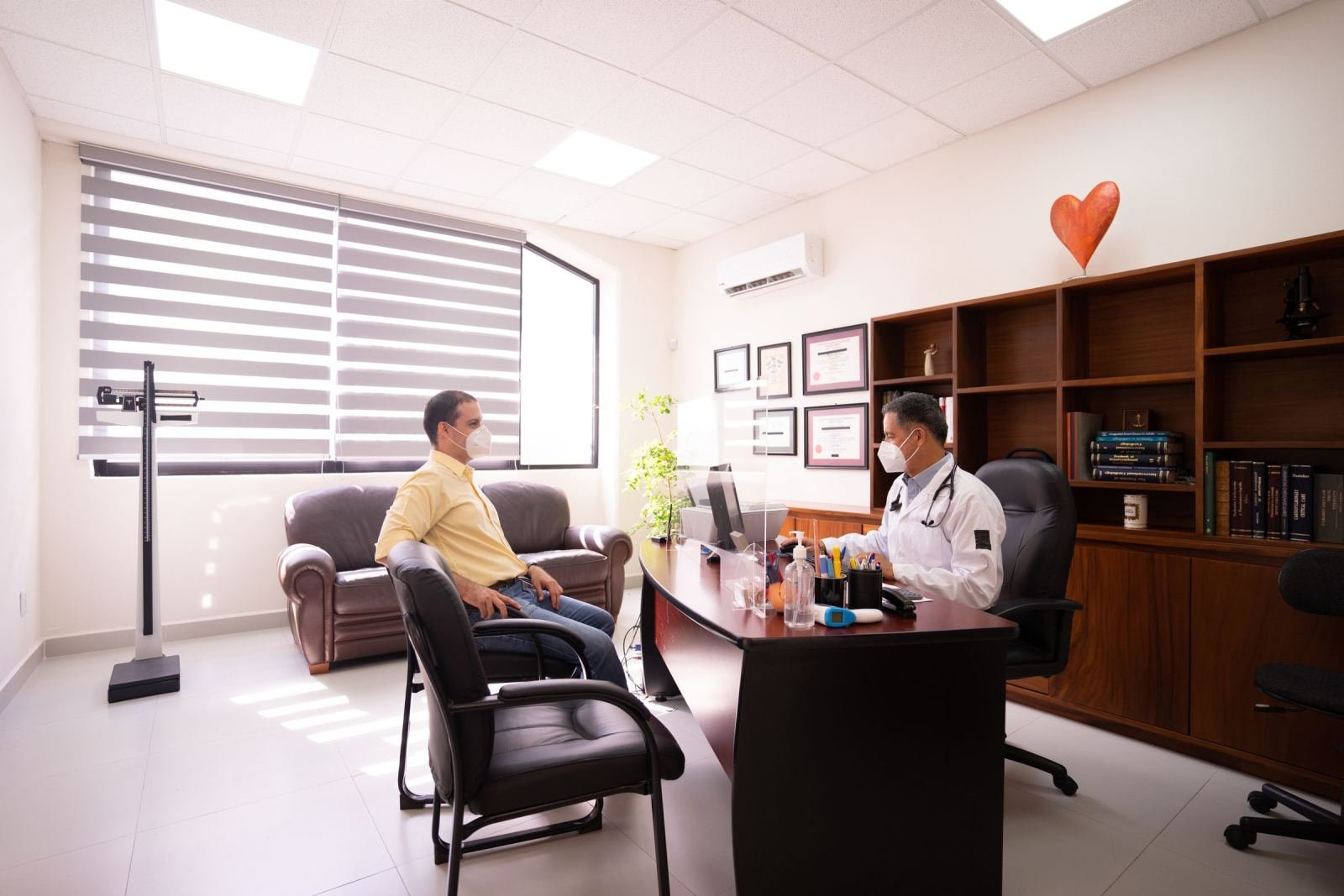 A masked doctor in a white coat chats with a patient in a bright room.