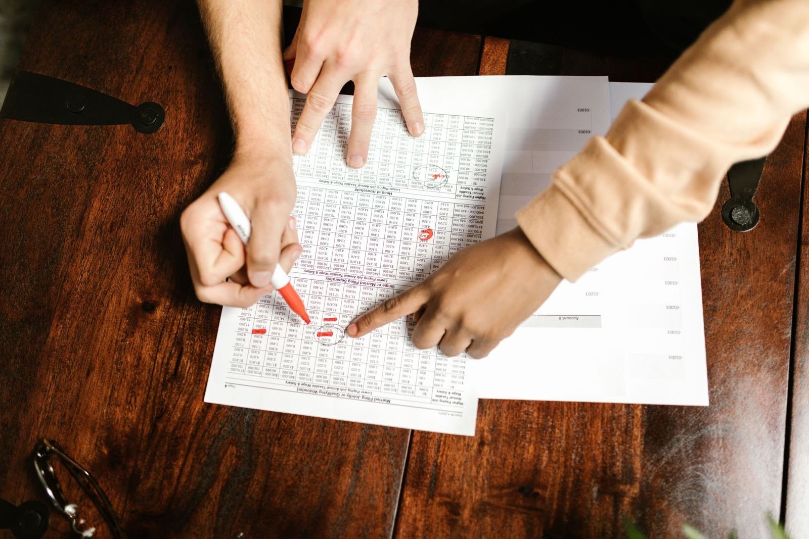 A pair of people mark up a paper fee list on a wood table using a red pen