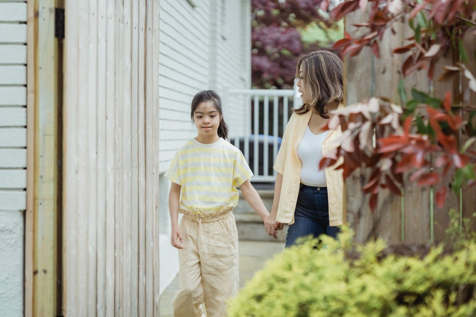 A woman and a young girl hold hands while walking outside next to a house