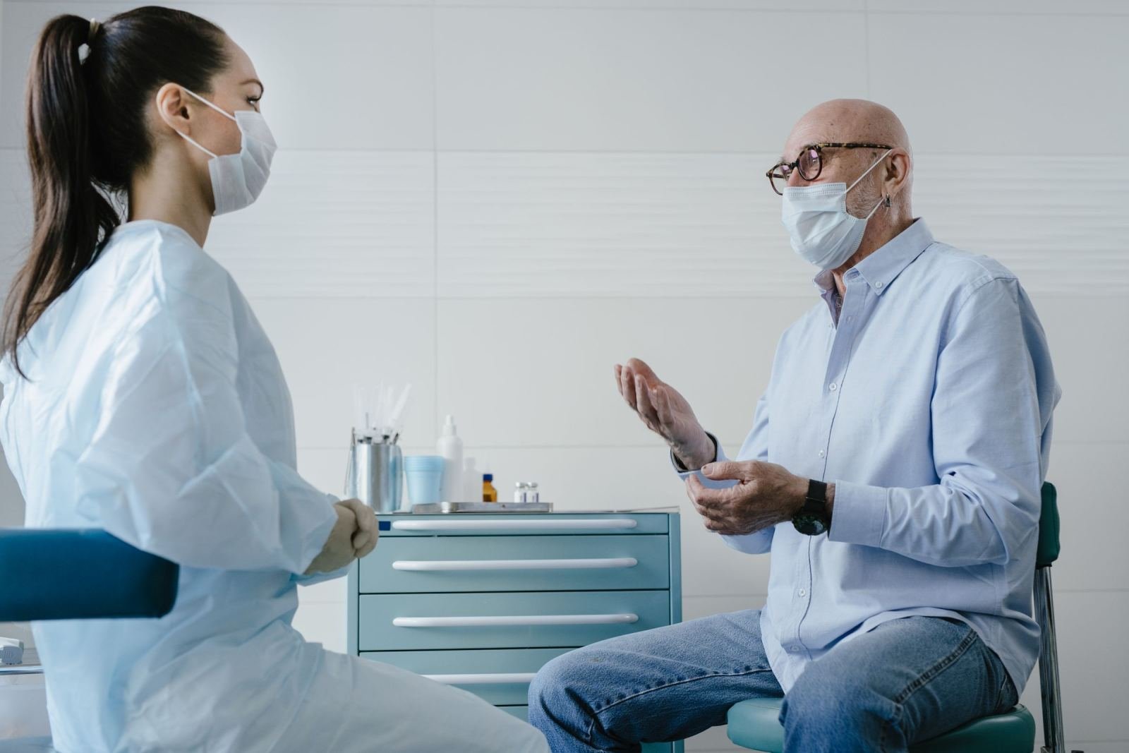 A masked healthcare worker in a gown talks to an older man who is also wearing a mask.