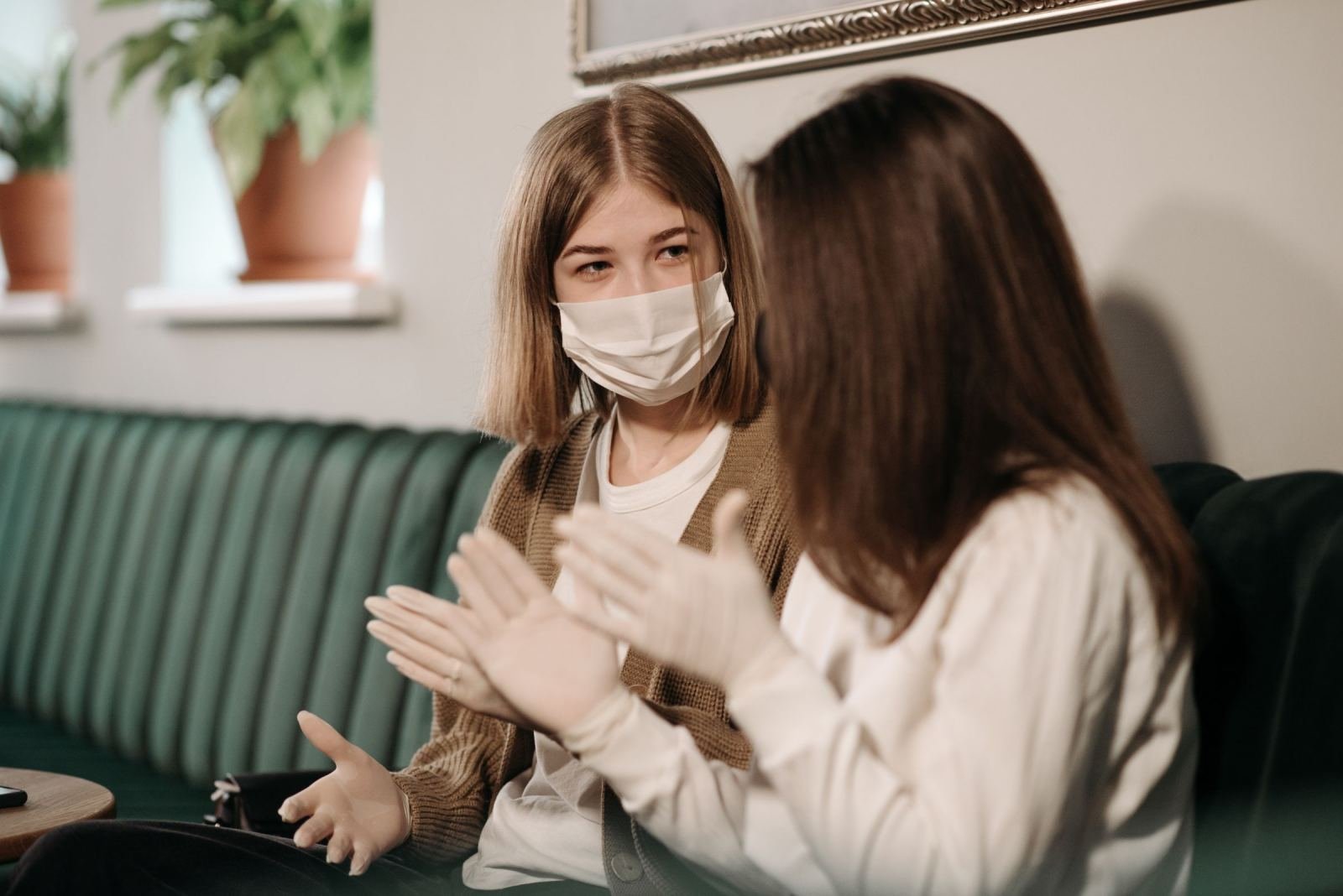 Two women talking on a green sofa, one wearing a white face mask