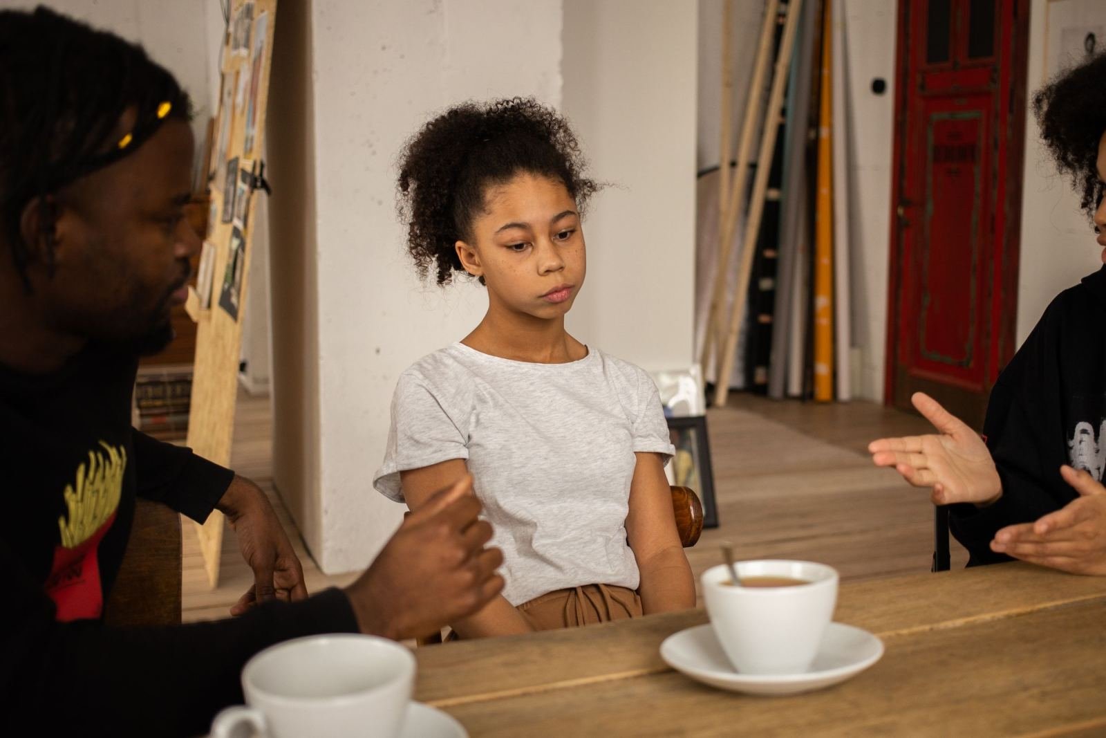 A young girl sits at a wooden table while a man and a woman talk with her.