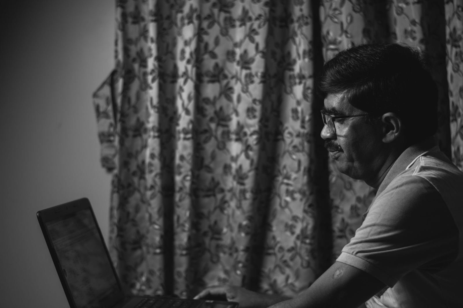 A man wearing glasses views a laptop screen while sitting before a patterned curtain