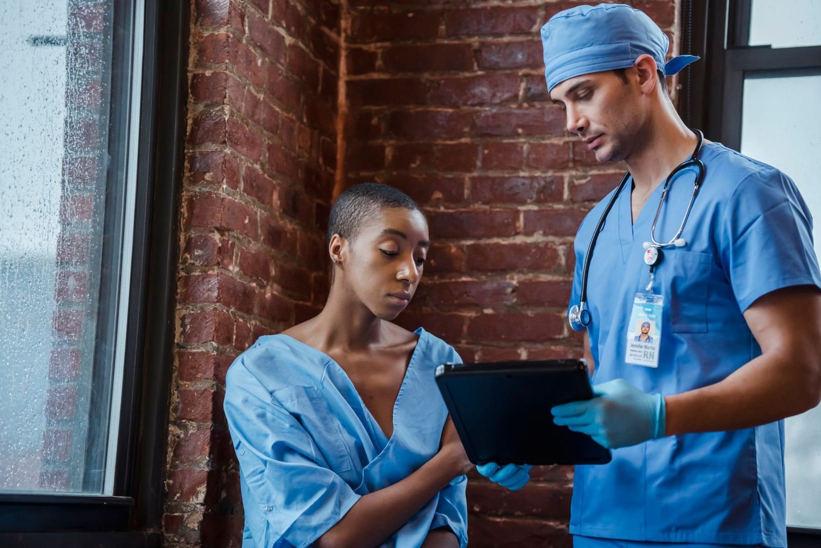 A patient in a blue gown looks at a tablet held by a nurse in blue scrubs