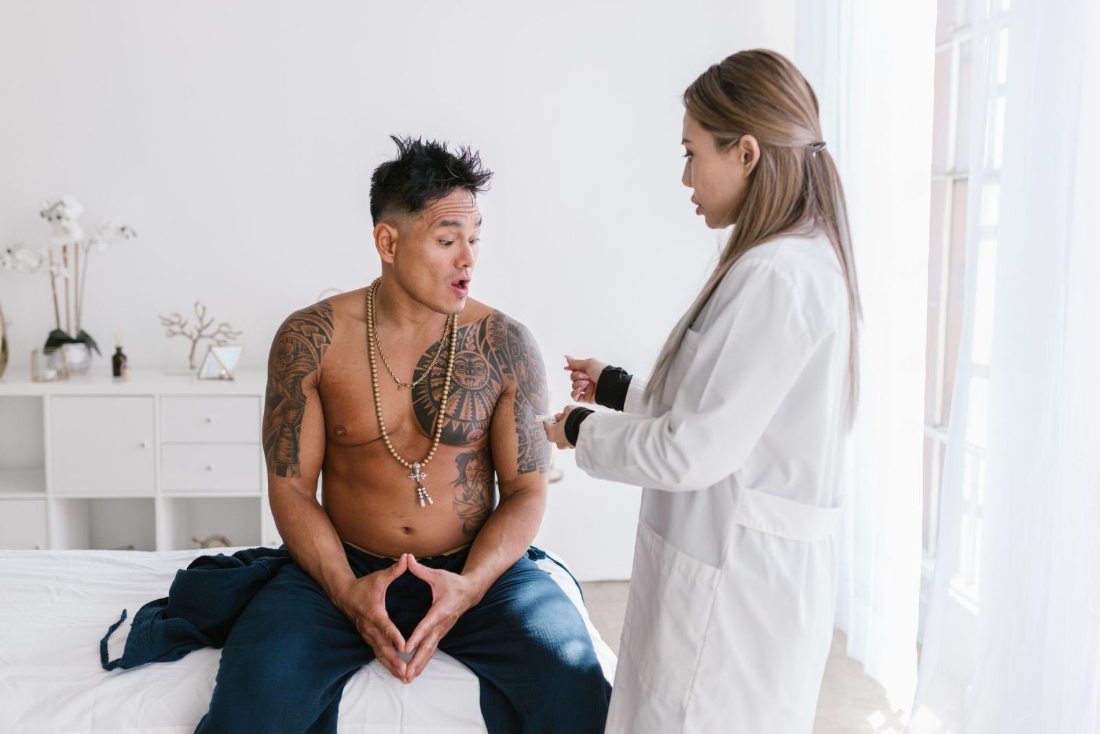 A shirtless man sits on an exam table while a clinician in a white coat talks to him.