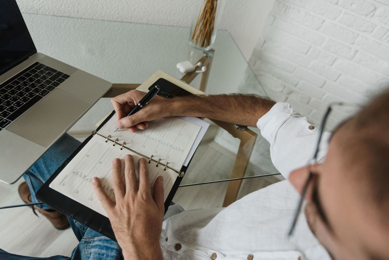 A person fills out a weekly planner on a glass desk near a laptop