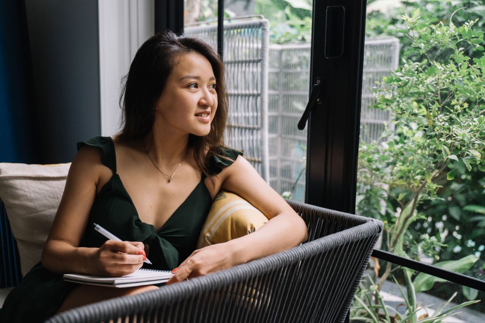 A woman in a green dress writes in a notebook while sitting by a window