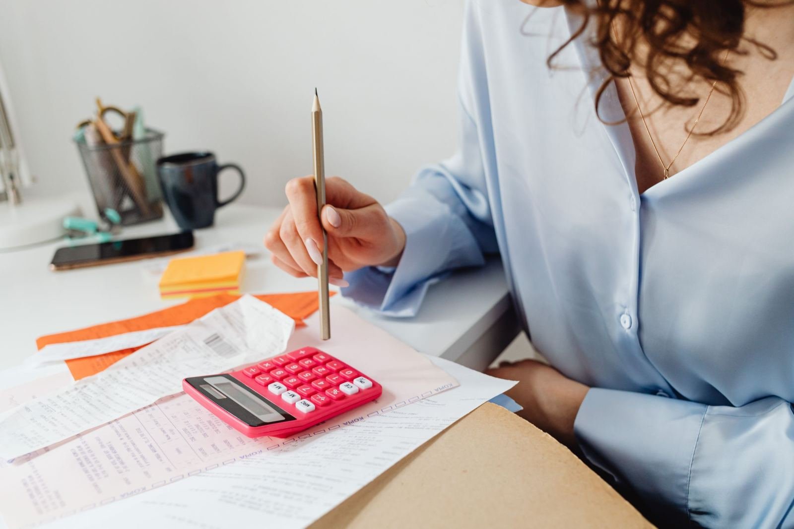 Someone sits at a desk, using a pink calculator and a pencil to go over papers.