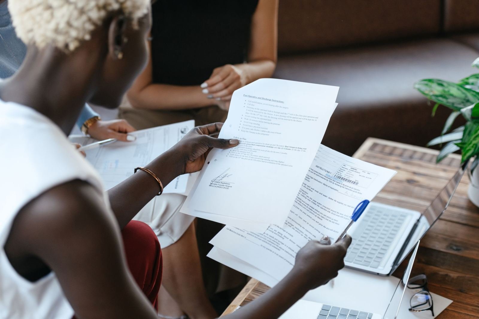 A woman sits at a table with a laptop, checking printed papers.