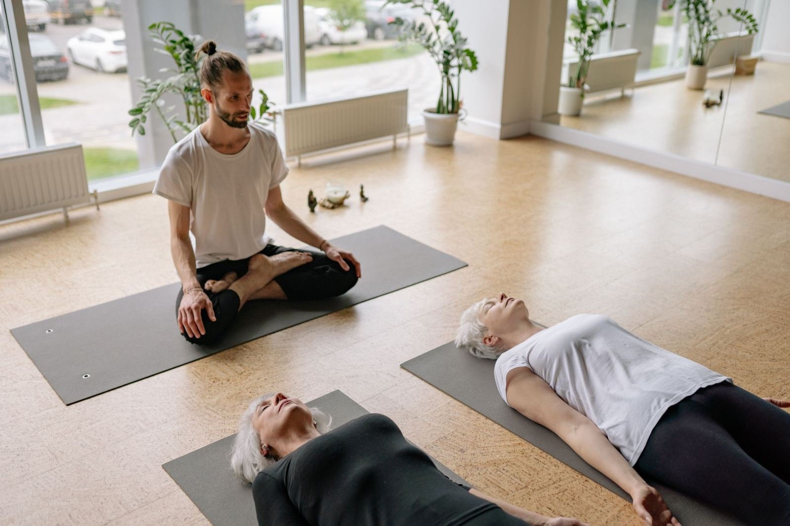 A man sits cross-legged on a mat facing two women lying on yoga mats in a bright studio
