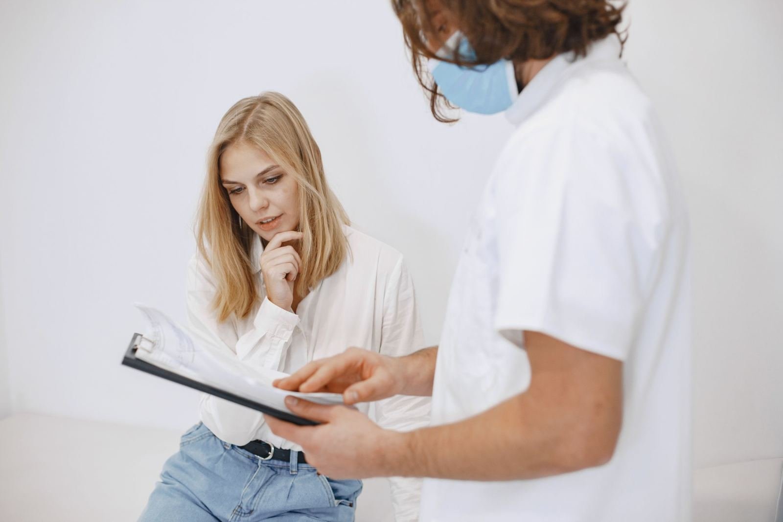 A masked doctor in a white coat displays a clipboard to a patient.