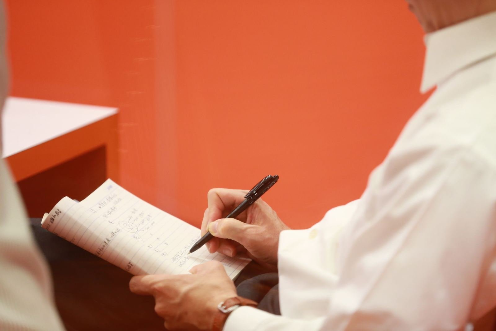 A person in a white shirt writing in a notebook against an orange wall