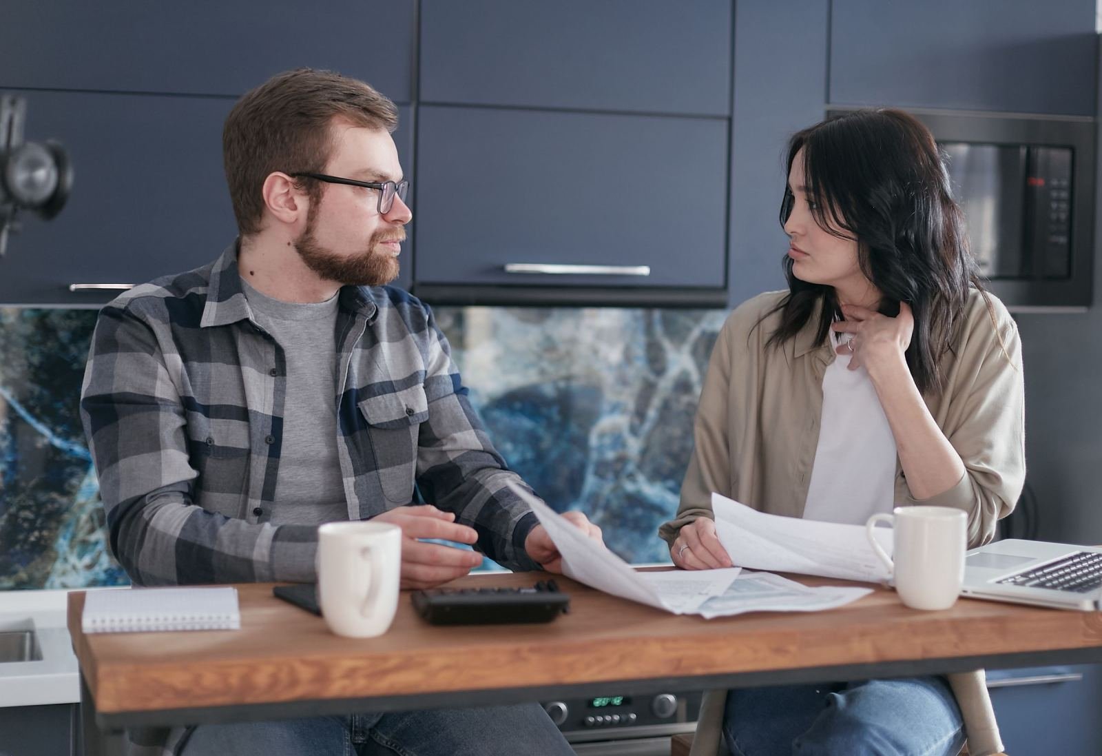 A couple sits at a kitchen table, looking over bills and using a calculator.