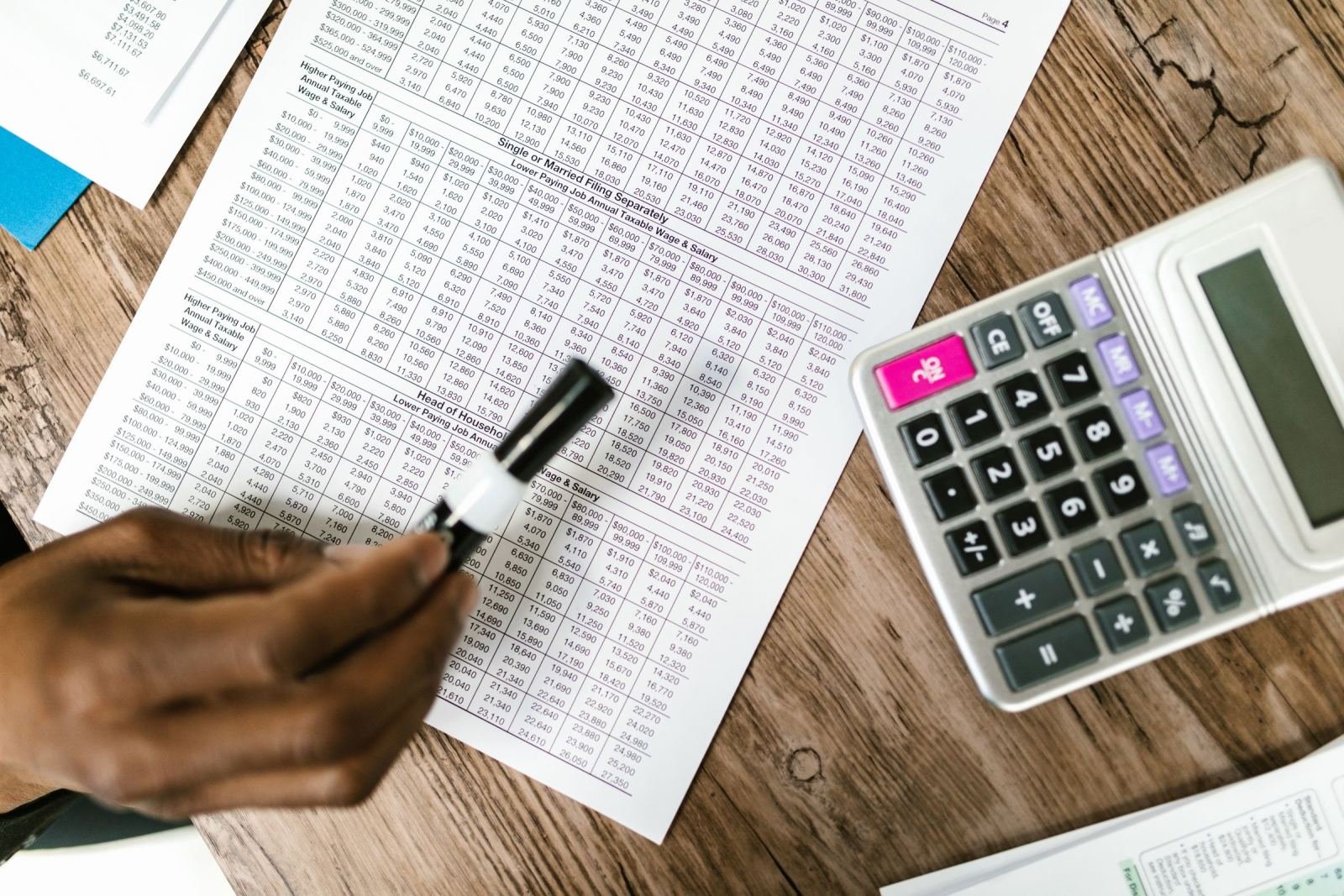 A hand holds a pen near a calculator on a wooden desk. A financial table sits next to them.